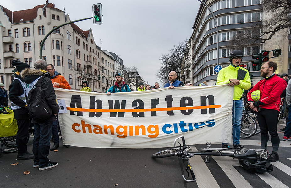 Mehrere Personen halten ein Banner auf dem das Wort "Abwarten" durchgestrichen ist. Sie Stehen auf einer städtischen Kreuzung.