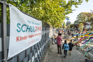 Kinder spielen auf der Straße. Ein Banner mit der Aufschrift "Schulzone - KInder haben Vorfahrt".