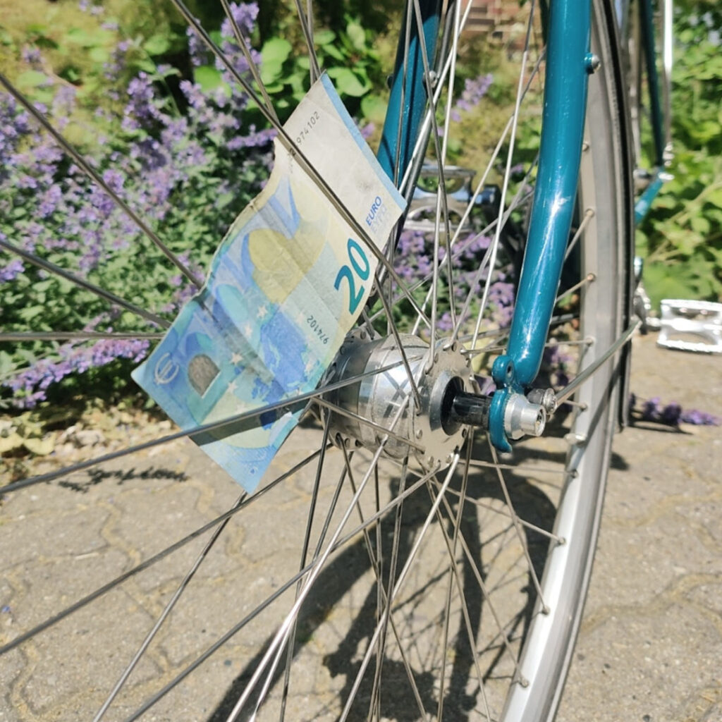 Foto eines Fahrrades. Es ist hauptsächlich das Vorderrad zu sehen, zwischen den Speichen steckt ein 20-Euro Schein. 