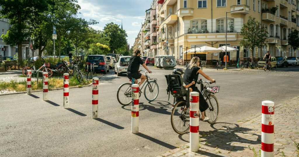 Foto einer Straßenkreuzung, im Vordergrund eine Reihe rot-weiß-gestreifter Poller, dazwischen fahren zwei Menschen Rad, kein Auto zu sehen, im Hintergrund Straßenbäume.