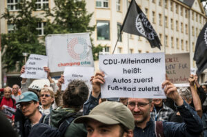 Protest auf der Straße: Eine Fahne mit Changing Cities, ein Schild mit der Aufschrift: CDU-Miteinander heißt: Autos zuerst, Fuß+Rad zuletzt.