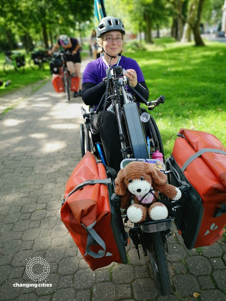 Sid, von vorne fotografiert, im Rollstuhl mit vorgespanntem Handbike. Vorn an dem Handbike sind ein Teddy und zwei Fahrradtaschen befestigt. Sid trägt einen grauen Fahrradhelm und ein lilanes T-Shirt.