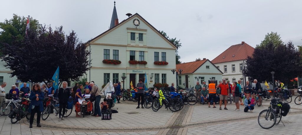 Panoramafoto: Duzende Menschen mit bepackten Fahrrädern vor einem gepflegten zweistöckigen weißen Haus mit der Aufschrift "Rathaus".