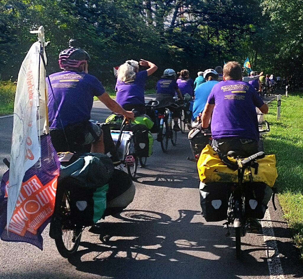 Duzende Radfahrende von hinten, viele tragel lila T-Shirts, alle haben Packtaschen, mehrere auch Fahnen an ihren Rädern