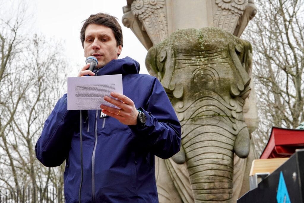 Ein Mann in blauer Jacke sprich in ein Mikro, hinter ihm ein großer steinerner Elefant, grauer Himmel.