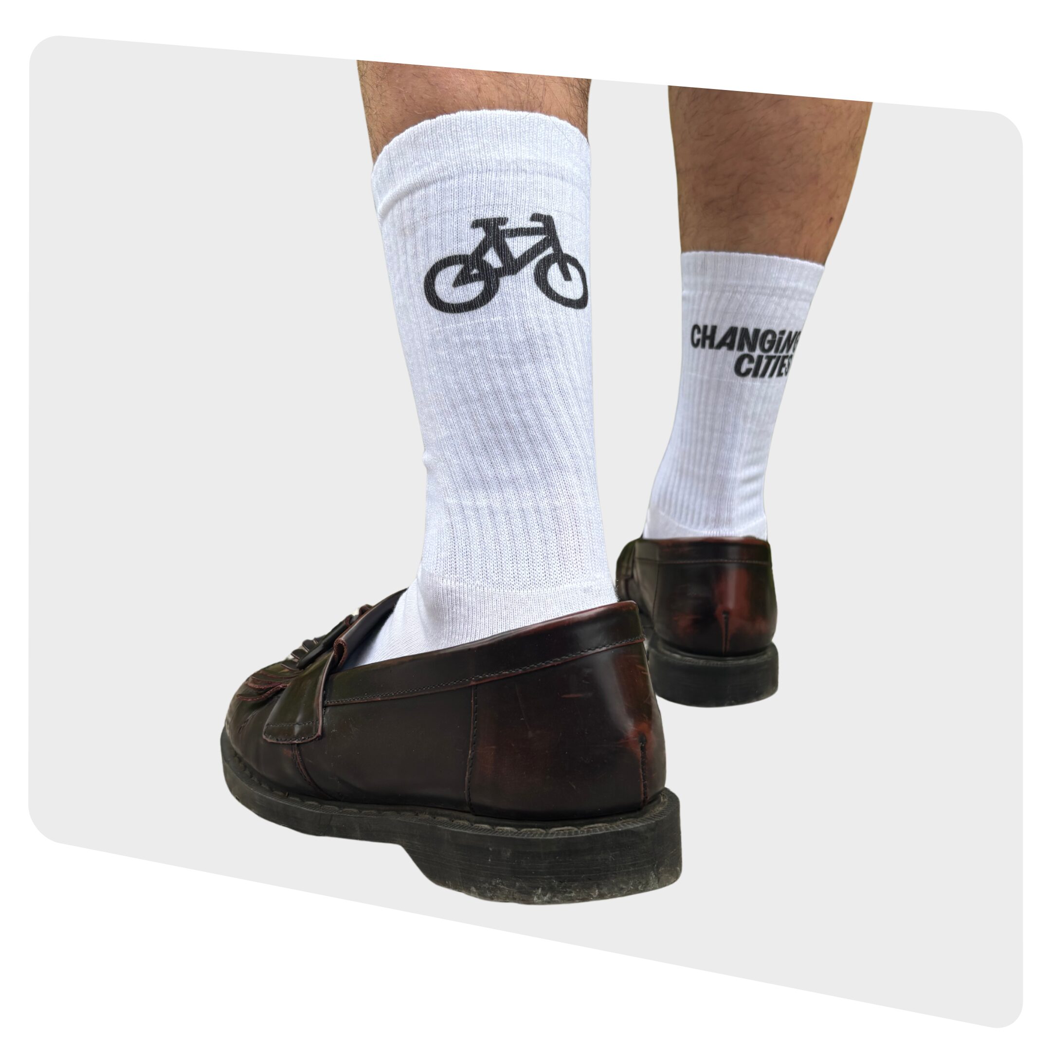 Nahaufnahme von Fahrradpedal, Hinterrad und Beinen mit weißen Socken auf pinkem Hintergrund. Auf der linken Socke ist das Logo von Changing Cities in schwarz gedruckt, auf der rechten ein Fahrradsymbol in Schwarz.