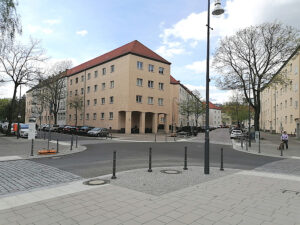 Ruhige Wohnstraße mit hellen Mehrfamilienhäusern, parkenden Autos, Bäumen und einem Straßenschild unter blauem Himmel. An den Straßenecken stehen etliche Poller.
