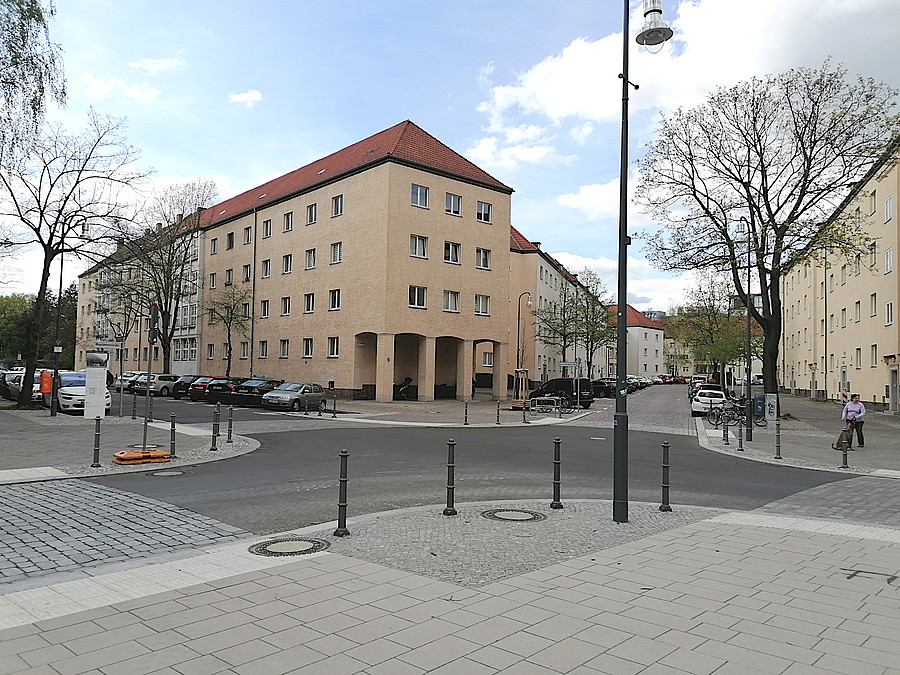 Ruhige Wohnstraße mit hellen Mehrfamilienhäusern, parkenden Autos, Bäumen und einem Straßenschild unter blauem Himmel. An den Straßenecken stehen etliche Poller.