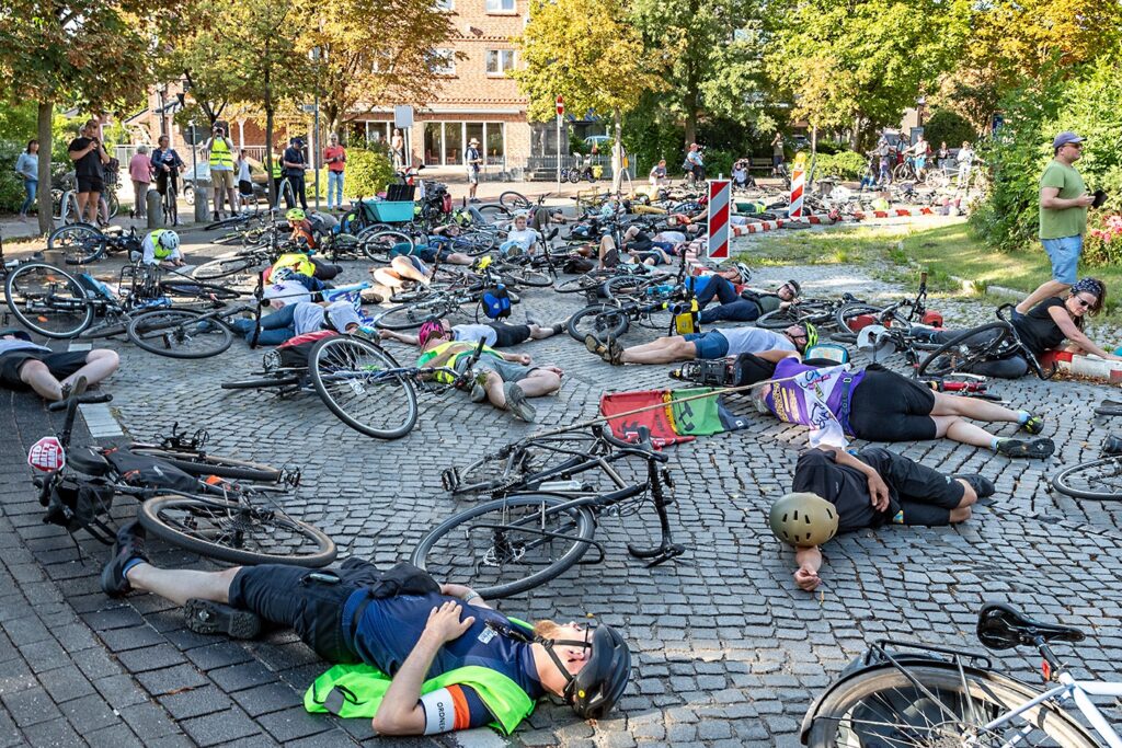 Radfahrer liegen wie angefahren auf einer Straße, während Fahrräder verstreut um sie herumliegen