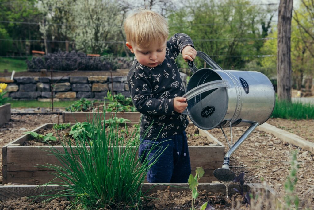 Kleinkind gießt Pflanzen im Garten mit einer Metallkanne