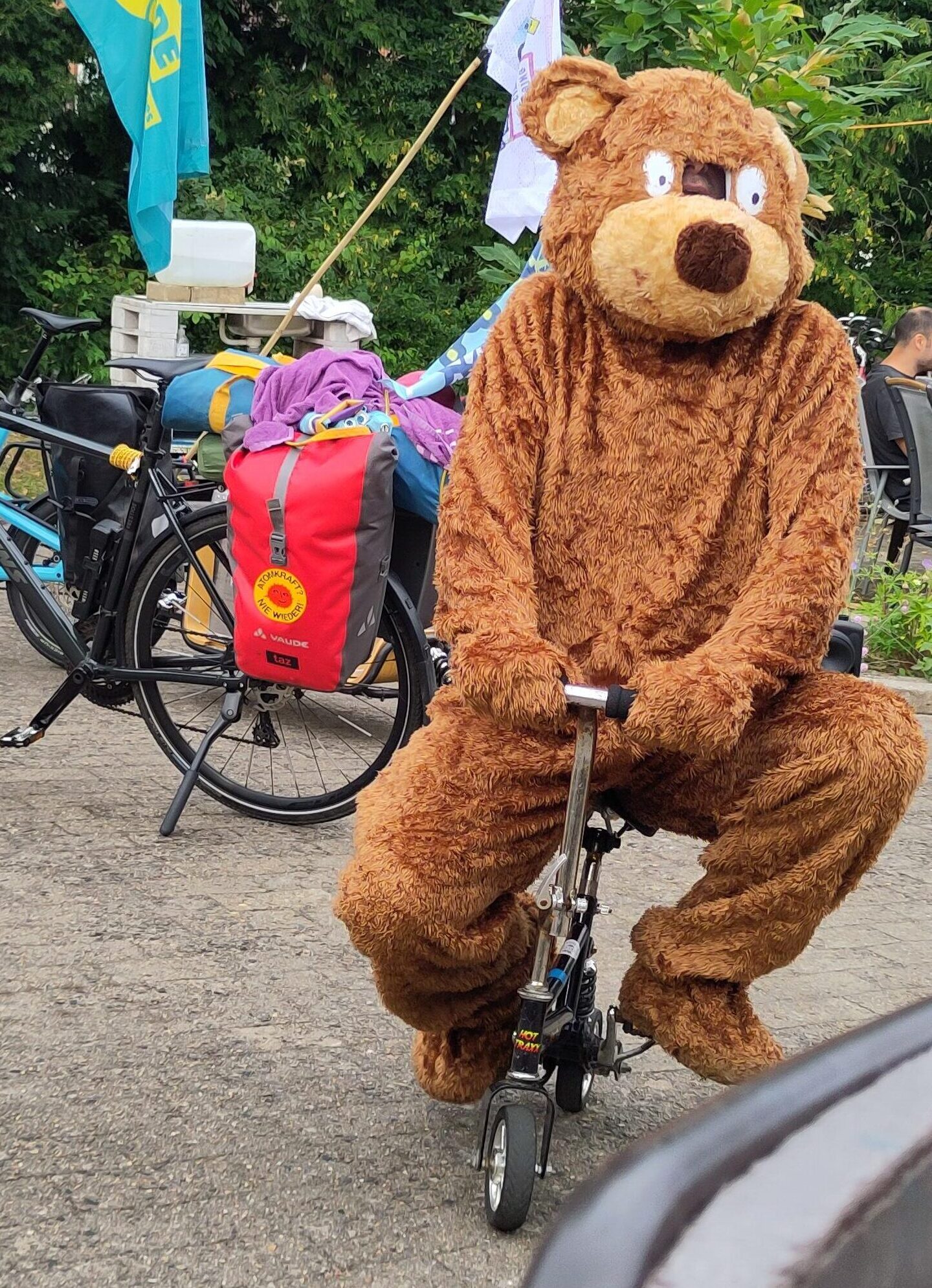 Bärenkostüm sitzt auf einem Mini-Rad vor Fahrrädern mit Bannern und einer Sitzgruppe im Camp