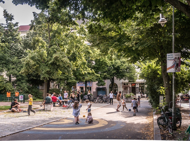 Kinder und Erwachsene treffen sich auf einem schattigen Platz; am Rand hängt ein Plakat zum „Berlin Circus Festival“